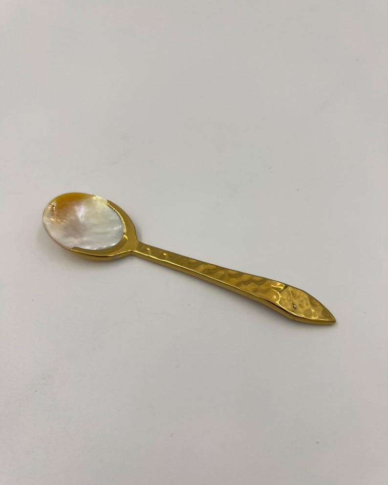 以下が含まれることがあります： 虹色に輝く白いマザーオブパールのボウルと、ハンマーで叩いたような質感のゴールドトーンの金属製ハンドルを備えた小さなスプーン。ハンドルは先端に向かって細くなっており、ボウルは金属製のフレームにしっかりと固定されています。