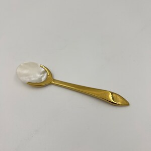 以下が含まれることがあります： 光沢のあるゴールドトーンの金属製ハンドルと、滑らかで虹色に輝く白いマザーオブパールのボウルを備えた小さなスプーン。ハンドルは先細りの尖った形状で、金属製のフレームが楕円形のシェルインサートをしっかりと固定しています。
