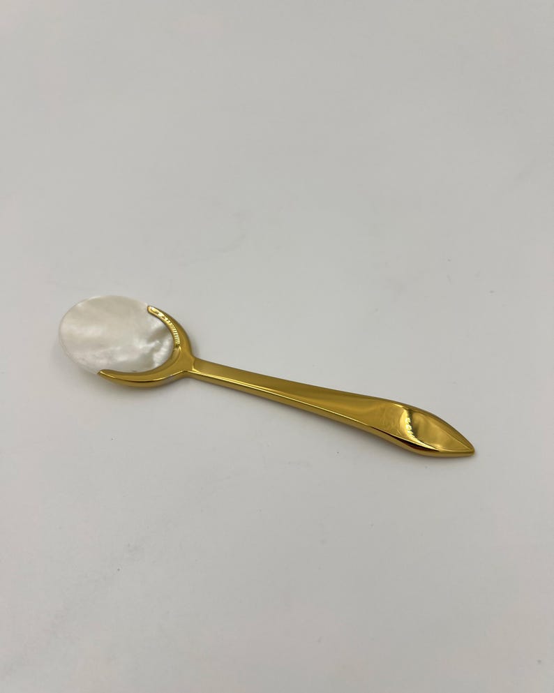 以下が含まれることがあります： 光沢のあるゴールドトーンの金属製ハンドルと、滑らかで虹色に輝く白いマザーオブパールのボウルを備えた小さなスプーン。ハンドルは先細りの尖った形状で、金属製のフレームが楕円形のシェルインサートをしっかりと固定しています。