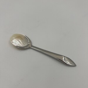 以下が含まれることがあります： 光沢のある銀色の金属製ハンドルと、虹色に輝く白いマザーオブパール製のボウルを備えた小さなスプーン。ハンドルは先端に向かって細くなっており、柄の端にはマザーオブパールのインレイが施されています。