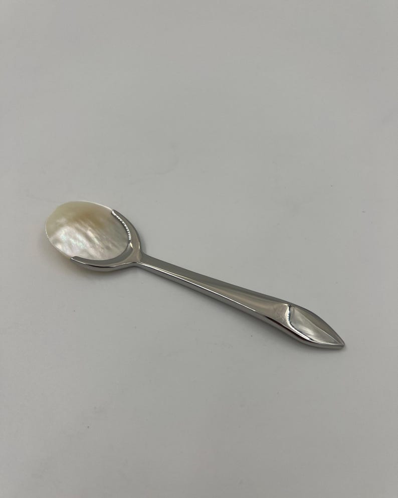 以下が含まれることがあります： 光沢のある銀色の金属製ハンドルと、虹色に輝く白いマザーオブパール製のボウルを備えた小さなスプーン。ハンドルは先端に向かって細くなっており、柄の端にはマザーオブパールのインレイが施されています。