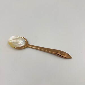 以下が含まれることがあります： 光沢のあるローズゴールドの金属製ハンドルと、虹色に輝く白いマザーオブパール（真珠母貝）のボウル部分を持つ小さなスプーン。スプーンは滑らかな曲線を描くデザインで、ハンドルの先端は尖っており、ボウル部分は丸みを帯びた光沢のある貝殻素材です。