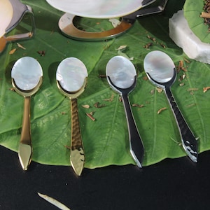May include: Four spoons with mother of pearl handles and different colored metal stems. The spoons are arranged on a green leaf.