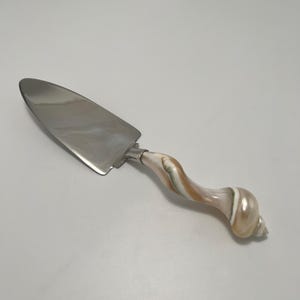 Puede incluir: Un servidor de pasteles con una hoja de metal plateado pulido y un mango hecho de una concha marina espiral e iridiscente con tonos arremolinados de crema, bronceado y blanco. El mango termina en una punta de concha natural.