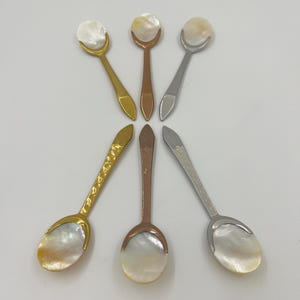 以下が含まれることがあります： 虹色に輝くマザーオブパール製のボウルと、ゴールド、カッパー、シルバーの色調の金属製ハンドルを備えた6本の小さなスプーンのセット。3本はプレーンなハンドルで、残りの3本は模様入りのテクスチャー加工されたハンドルで、先端が尖った形状をしています。