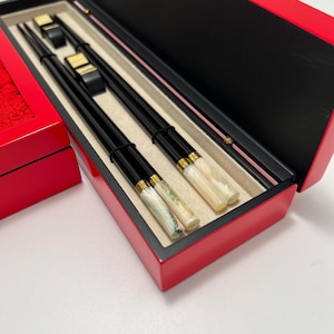 Op de afbeelding: Een paar zwarte eetstokjes met iriserende, parelmoerachtige uiteinden en goudkleurige banden, rustend in een rechthoekige rode opbergdoos met een beige binnenvoering. Twee kleine zwarte en goudkleurige eetstokjessteunen zijn inbegrepen.