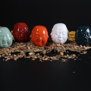 Puede incluir: Cinco figuritas de cerámica de caras sonrientes y frunciendo el ceño, pintadas en diferentes colores, incluyendo verde, rojo, naranja, blanco y negro. Las figuritas están dispuestas sobre una cama de virutas de madera.