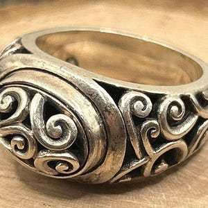 May include: A silver ring with a detailed, swirling design. The ring is wide and has a rounded top.