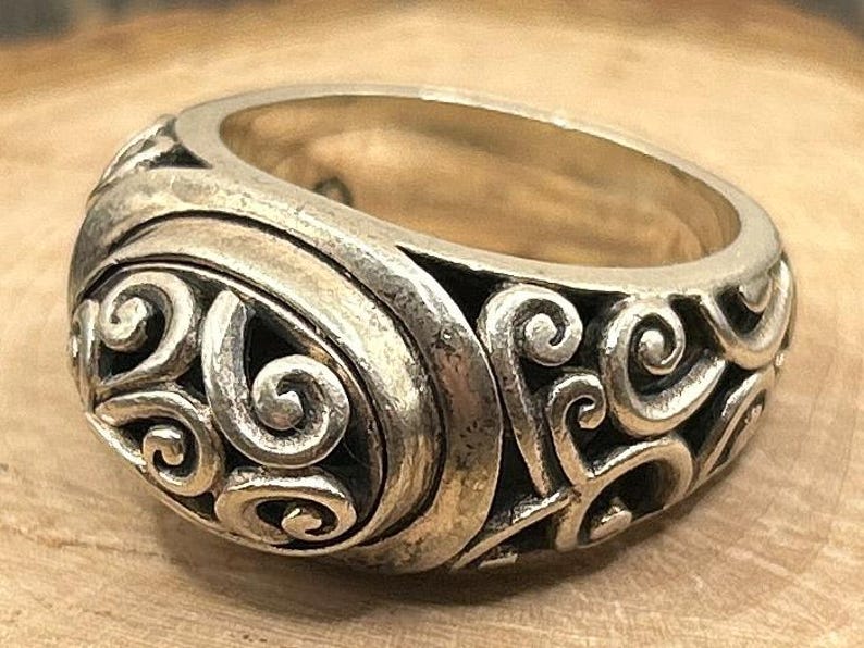 May include: A silver ring with a detailed, swirling design. The ring is wide and has a rounded top.