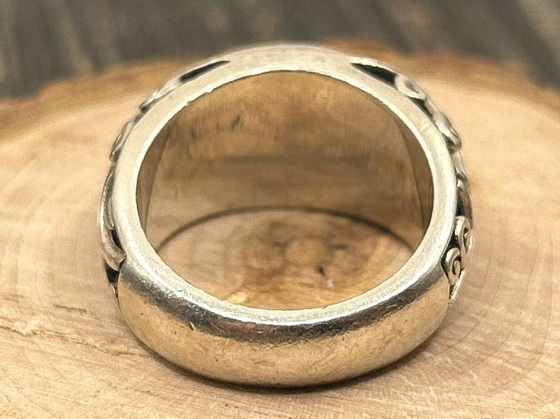 May include: A silver ring with intricate designs on the band. The ring is wide and has a smooth, rounded surface.