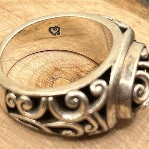 May include: A silver ring with a detailed, ornate design. The ring has a heart-shaped marking on the inside.