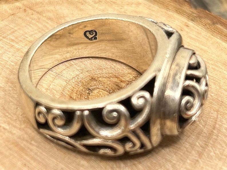 May include: A silver ring with a detailed, ornate design. The ring has a heart-shaped marking on the inside.