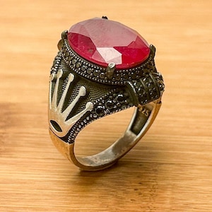 Könnte beinhalten: Ein silberner Ring mit einem großen roten Edelstein und einem Kronendesign. Der Ring ist mit schwarzen Steinen um den Edelstein besetzt.
