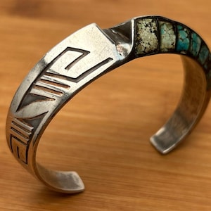 May include: Silver cuff bracelet featuring geometric patterns and turquoise inlay. The bracelet is curved and set against a wooden background. The turquoise stones are a mix of blue and green.