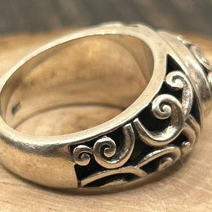 May include: A silver ring with a detailed, swirling design. The ring is wide and has a rounded top.
