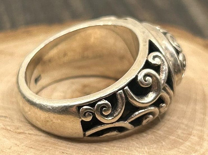 May include: A silver ring with a detailed, swirling design. The ring is wide and has a rounded top.