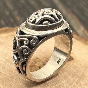 May include: A silver ring with an intricate design of swirling lines and a raised oval center. The ring is on a wooden surface.