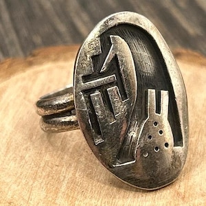 Könnte beinhalten: Ein silberfarbener Ring mit einem ovalen Gesicht, das ein abstraktes Design zeigt. Die Oberfläche des Rings ist strukturiert und weist ein erhabenes Muster auf. Das Band hat ein Doppelband-Design. Der Ring ist auf einer Holzoberfläche platziert.