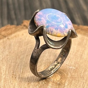 May include: A vintage sterling silver ring featuring a large, oval opal gemstone. The opal displays a play of color with hues of blue, pink, and yellow. The ring has a unique, sculptural design with the hallmark "STERLING" and "925" visible.