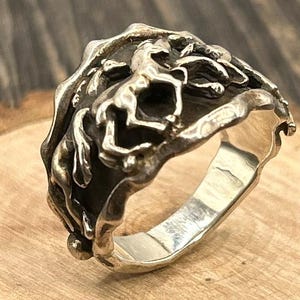 May include: A silver ring with a detailed design featuring a horse and foliage. The ring has a textured, organic appearance with a polished inner band. The horse is depicted in a dynamic pose, adding a sense of movement to the piece.