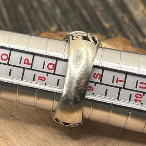 May include: A silver ring with an intricate design, measured on a ring sizer. The ring size is 9.