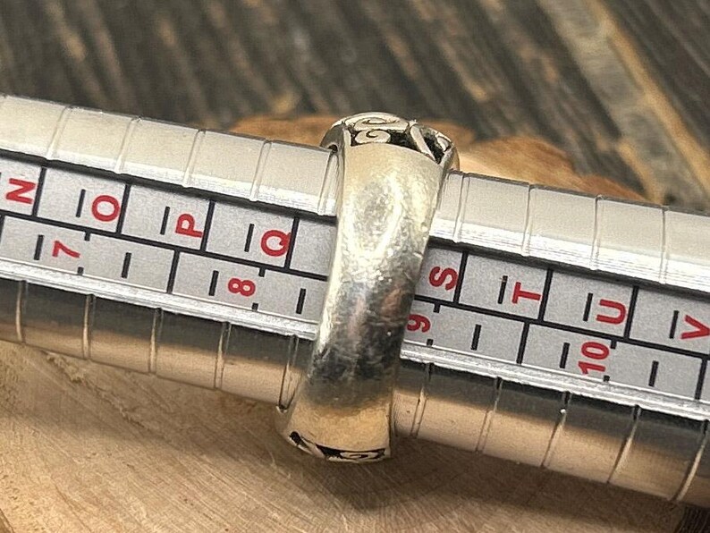 May include: A silver ring with an intricate design, measured on a ring sizer. The ring size is 9.