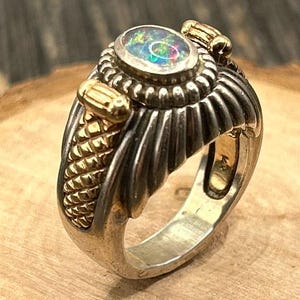 May include: A silver and gold ring with a large opal gemstone set in the center. The ring has a textured band with a braided design.