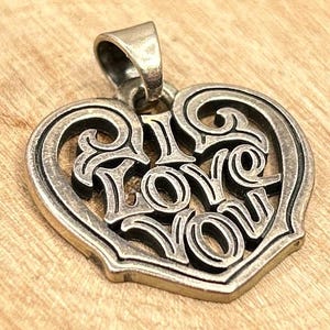 May include: Silver-toned heart-shaped pendant with the words "I LOVE YOU" in a stylized font. The pendant has an ornate, openwork design and a bail for attaching to a chain. The pendant is displayed on a wooden surface.