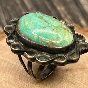 May include: Oval turquoise ring with a silver-tone setting. The turquoise stone has a mottled green and brown pattern. The ring has a decorative, scalloped bezel and a split shank design.