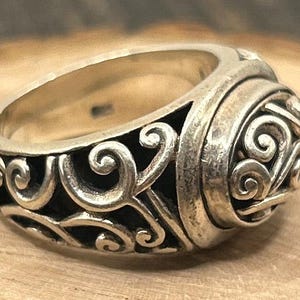 May include: A silver ring with a detailed, ornate design featuring swirling patterns. The ring is wide and has a rounded top.