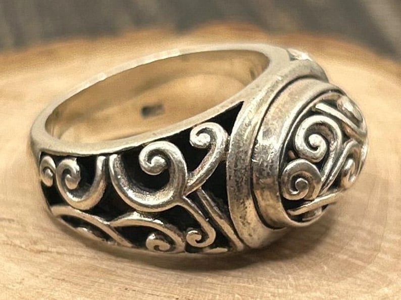 May include: A silver ring with a detailed, ornate design featuring swirling patterns. The ring is wide and has a rounded top.