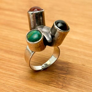May include: A silver ring with three stones: a green stone, a red stone, and a black stone. The ring is abstract and modern in design.