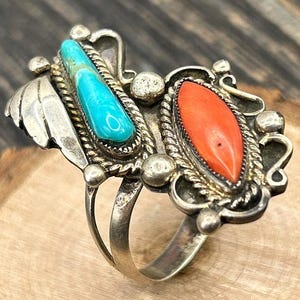 May include: A silver ring with two large, oval-shaped stones. One is a vibrant turquoise, the other a bright orange. The ring has intricate silver detailing, including a leaf-like design and small silver beads. The ring is likely a piece of Native American jewelry.