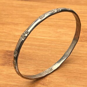 May include: A silver-colored bangle bracelet with a floral pattern. The bracelet is made of a shiny metal and has a slightly textured surface. The floral design is repeated around the entire bracelet.