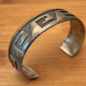 May include: A silver-toned cuff bracelet with a geometric pattern. The open cuff design features a series of repeating angular shapes, creating a textured surface. The bracelet is displayed on a wooden surface.