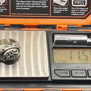 May include: A silver ring with an intricate design is placed on a digital scale. The scale reads 1.44.59. The scale is black with a silver display and buttons labeled "TARE", "ON/OFF", and "MODE".