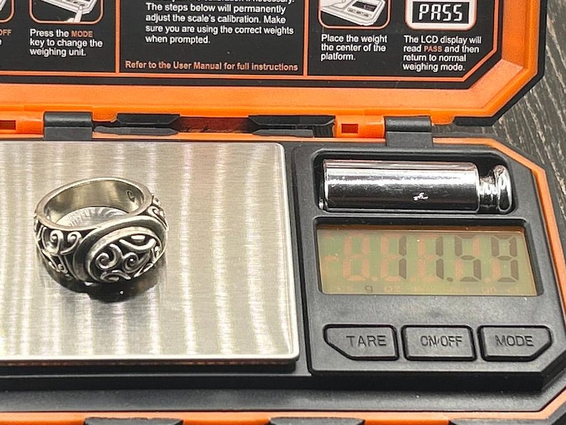 May include: A silver ring with an intricate design is placed on a digital scale. The scale reads 1.44.59. The scale is black with a silver display and buttons labeled "TARE", "ON/OFF", and "MODE".