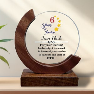 Puede incluir: Una placa de madera marrón con un círculo de vidrio transparente en el centro. El círculo de vidrio tiene un diseño de estrellas doradas y el texto "6 Years of Service Jean Fluck For your teaching leadership & teamwork In honor of your service to patients and staff at BTH".