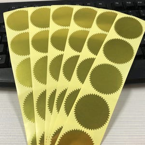 Op de afbeelding: Een vel met gouden folie stickers met een gegolfde rand. De stickers zijn in rijen gerangschikt en zijn klaar om te worden gebruikt voor het verzegelen van enveloppen, het toevoegen van een decoratieve toets aan knutselwerk of voor andere creatieve projecten.