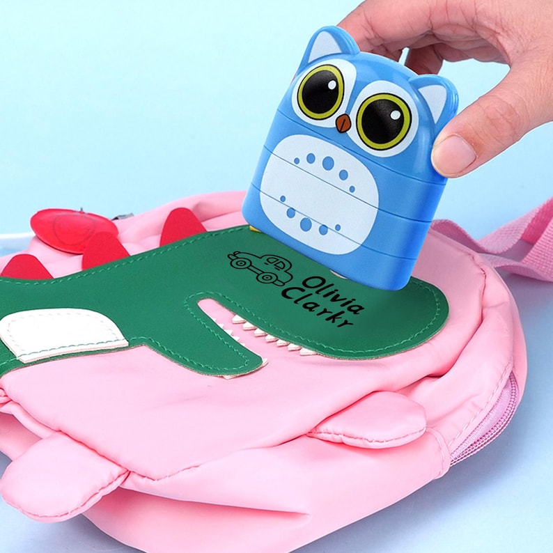 May include: A blue owl-shaped stamp with large eyes and a white belly is being placed on a pink backpack with a green alligator design. The stamp has the text "Olivia Clarkr" on it.