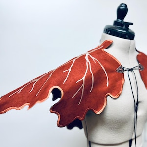 Puede incluir: Una pieza de hombro de gamuza roja en forma de hoja con costuras blancas y un cordón negro. La pieza se muestra en un maniquí blanco.