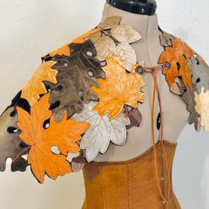 May include: A leather shoulder cape featuring a leaf design in shades of orange, brown, and beige. The cape is adorned with leaf cutouts and secured with a leather tie and decorative buttons. The cape is displayed on a mannequin.