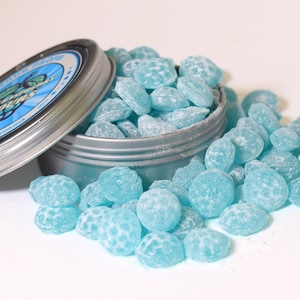 May include: A silver tin with a blue and white label, partially open, spilling out numerous light blue candies. The candies have a textured surface and are scattered around the tin. The label reads "Sweet Paws" with a butterfly graphic.