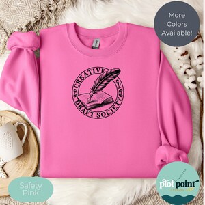 Puede incluir: Una sudadera rosa con un diseño negro de pluma y libro abierto, rodeado por las palabras "CREATIVE DRAFT SOCIETY". La sudadera es de color rosa de seguridad sólido, con el texto "More Colors Available!" en un círculo gris oscuro.