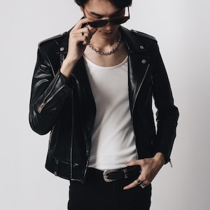 Könnte beinhalten: Eine schwarze Leder-Motorradjacke mit silberfarbenen Metallreißverschlüssen und Druckknöpfen, getragen über einem schlichten weißen Unterhemd. Die Jacke verfügt über Schulterklappen, Reißverschlussbündchen und einen Reverskragen, kombiniert mit einer schwarzen Hose und einem Gürtel.