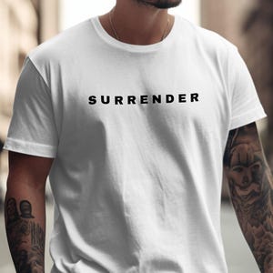 May include: White t-shirt with the word "SURRENDER" printed in black across the chest. The shirt has short sleeves and a crew neck. The person wearing the shirt has tattoos on their arms.