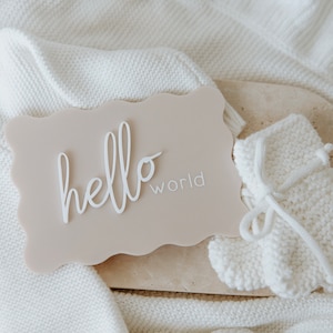 May include: A light brown sign with the words "hello world" in white lettering. The sign is on a white knitted blanket with a white knitted baby bootie.
