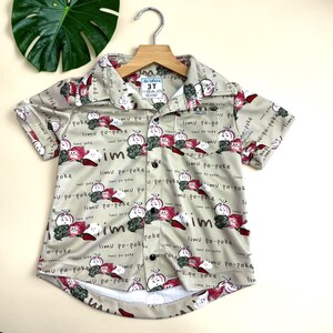 Può includere: Una camicia a maniche corte di colore beige chiaro, con motivi di illustrazioni di verdure in stile cartone animato e le parole "limu po-poke". La camicia ha una piccola tasca ed è etichettata "3T".