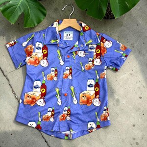 Può includere: Una camicia azzurra a maniche corte con un motivo ripetuto di gatti arancioni, bottiglie e cipolle. La camicia ha una piccola tasca e un colletto. L'etichetta recita "3T".