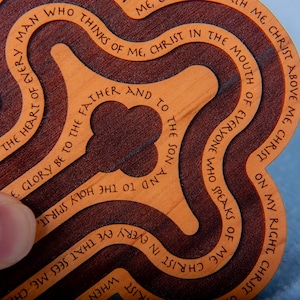 May include: A wooden cross-shaped item with a layered design. The cross features engraved text in a circular pattern, with a dark brown outer layer and a lighter brown inner layer. The text is in English.